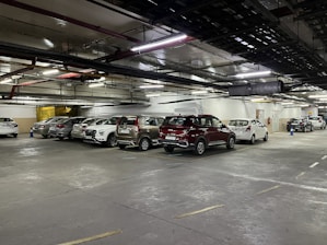 A dimly lit underground parking lot with numerous parked cars in designated spaces. The ceiling has visible pipes and ventilation ducts, and there are large overhead lights illuminating the area. Blue traffic cones are placed intermittently.