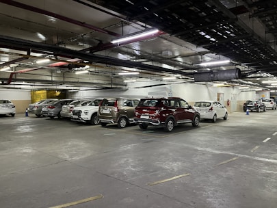 A dimly lit underground parking lot with numerous parked cars in designated spaces. The ceiling has visible pipes and ventilation ducts, and there are large overhead lights illuminating the area. Blue traffic cones are placed intermittently.