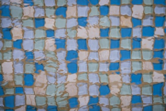 A pattern of blue, green, and beige tiles is visible through the rippling surface of water. The tiles create a grid-like layout, with the water distorting and reflecting the shapes and colors, giving an impressionistic effect.