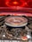 A close-up view of a car engine with a chrome air filter cover that has a checkered decal on top. The surrounding components include various pipes, cables, and bolts. The hood of the car, painted in a shiny red with a reflective finish, is partially visible in the background, casting an orange glow over the engine parts.