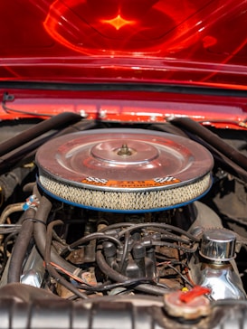A close-up view of a car engine with a chrome air filter cover that has a checkered decal on top. The surrounding components include various pipes, cables, and bolts. The hood of the car, painted in a shiny red with a reflective finish, is partially visible in the background, casting an orange glow over the engine parts.