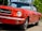 A red classic car with a black convertible top is parked on a roadway. The car's shiny chrome details and emblematic badges are clearly visible, indicating that it is well-maintained. The background includes lush green foliage and a bright sky, suggesting it is daytime.