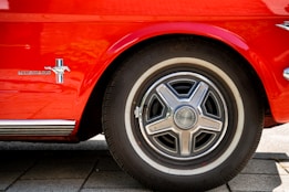 A close-up view of a classic car's wheel and logo, showcasing the shiny chrome hubcap on a black tire with white sidewall. The red car body has a 'Mustang' logo with a galloping horse emblem against a striped background.