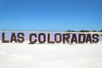 A large sign reading 'Las Coloradas' stands prominently on a sandy, white ground in front of a pastel pink body of water. The sky above is clear and blue, indicating a sunny day. In the distance, there is a line of greenery along the horizon.