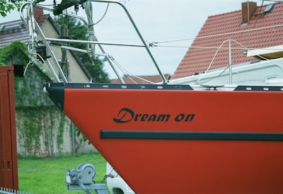 A red sailboat with the words 'Dream on' written on the side is positioned on a trailer. The boat is in a grassy area with red-roofed houses in the background and some greenery visible.