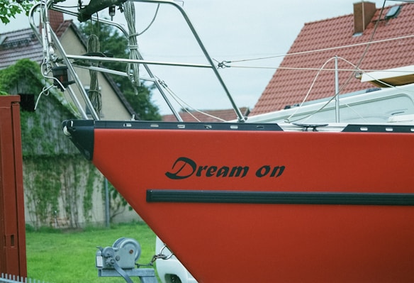 A red sailboat with the words 'Dream on' written on the side is positioned on a trailer. The boat is in a grassy area with red-roofed houses in the background and some greenery visible.