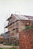 A weathered, old building is surrounded by scaffolding, indicating it is under restoration or construction. The building has a sloped roof with visible wear, and the exterior walls show signs of age and peeling. A brick wall is in the foreground, adding texture and depth to the scene.