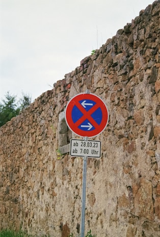 A weathered stone wall with a no parking traffic sign featuring arrows pointing left and right. The sign includes additional text indicating restrictions starting from a specific date and time in German. The background features some greenery.