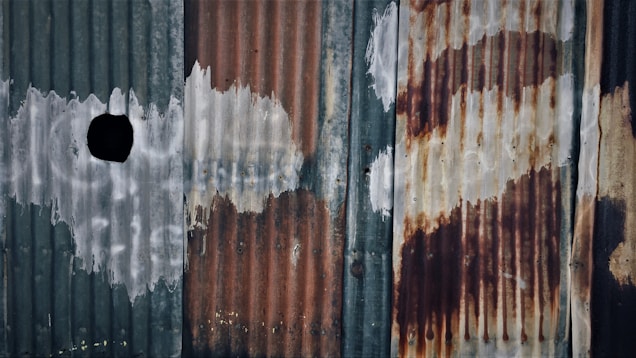 Corrugated metal sheets with a distressed and weathered appearance, showing signs of rust and peeling paint. There are streaks and blotches of white and reddish-brown across the surface, with a significant black hole or mark on the left side.