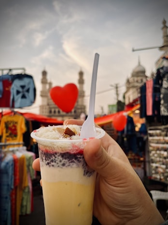 A hand holds a dessert cup filled with multiple layers, including yellow, white, and red with a topping of what appears to be nuts and grated coconut. A plastic spoon is placed vertically in the cup. The background shows a bustling market setting with clothes on display and a red heart-shaped decoration. A historic building with minarets is visible in the distance under a partly cloudy sky.