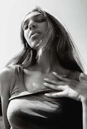 A black and white photograph of a person with long hair, looking up towards the camera. The perspective is from a low angle, emphasizing the person's facial features and expression. The person is wearing a sleeveless top and has one hand touching their chest. The lighting creates strong contrasts, highlighting the contours of the face and hair.