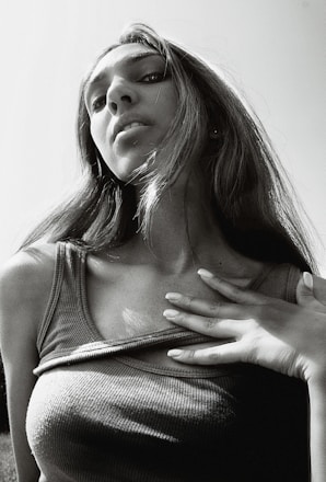 A black and white photograph of a person with long hair, looking up towards the camera. The perspective is from a low angle, emphasizing the person's facial features and expression. The person is wearing a sleeveless top and has one hand touching their chest. The lighting creates strong contrasts, highlighting the contours of the face and hair.