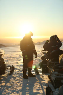 Sunset silhouette of a rider standing on a ridge, snowscoot beside them, overlooking the alpine landscape.