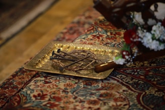 A decorative metal tray, possibly made of brass or a similar material, placed on an intricately patterned carpet. The tray contains several long, thin branches or twigs arranged in a circular pattern. Next to the tray, there are some vibrant red and white flowers, possibly part of a floral arrangement.