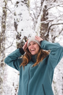 A smiling person wearing a chunky knit hat, walking through a snowy park with bare trees.
