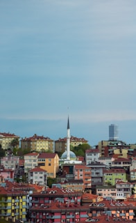 A landscape showcasing a densely populated urban area with colorful residential buildings and a prominent mosque with a tall minaret in the center. The skyline includes a modern skyscraper in the background against a bright blue sky.