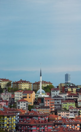 A landscape showcasing a densely populated urban area with colorful residential buildings and a prominent mosque with a tall minaret in the center. The skyline includes a modern skyscraper in the background against a bright blue sky.