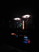 A computer setup is illuminated with colorful LED lights. The components, such as cooling fans with white light rings and RGB elements, create a vibrant and high-tech atmosphere. Various hardware brands are visible, with emphasis on aesthetic lighting.