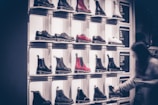 A display wall features multiple pairs of boots arranged on shelves, with variations in color including black, white, and red. Each boot is notably chunky with a thick sole. A blurred figure of a person is seen on the right side, likely examining the footwear.