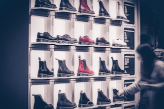 A display wall features multiple pairs of boots arranged on shelves, with variations in color including black, white, and red. Each boot is notably chunky with a thick sole. A blurred figure of a person is seen on the right side, likely examining the footwear.