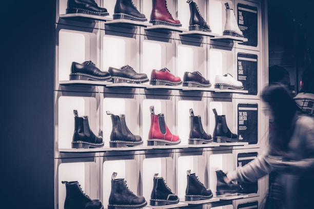 A display wall features multiple pairs of boots arranged on shelves, with variations in color including black, white, and red. Each boot is notably chunky with a thick sole. A blurred figure of a person is seen on the right side, likely examining the footwear.