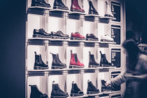 A display wall features multiple pairs of boots arranged on shelves, with variations in color including black, white, and red. Each boot is notably chunky with a thick sole. A blurred figure of a person is seen on the right side, likely examining the footwear.