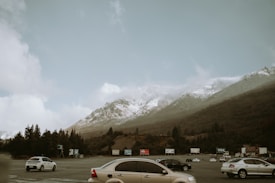 Snow-capped mountains rise in the background under a cloudy sky, with lush green forests at their base. In the foreground, a parking lot with several vehicles including sedans and hatchbacks is visible. Advertising billboards are positioned at the edge of the lot.