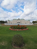 An elegant historic palace sits prominently in the center, featuring a symmetrical design with intricate architectural details. Lush green lawns surround the structure, adorned with meticulously arranged flowerbeds bursting with vibrant red and white blooms. A manicured hedge in the foreground adds to the garden's charm. Above, a partly cloudy sky offers a serene backdrop.