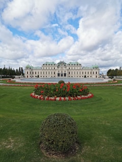 An elegant historic palace sits prominently in the center, featuring a symmetrical design with intricate architectural details. Lush green lawns surround the structure, adorned with meticulously arranged flowerbeds bursting with vibrant red and white blooms. A manicured hedge in the foreground adds to the garden's charm. Above, a partly cloudy sky offers a serene backdrop.
