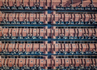 An aerial view reveals a structured layout of numerous closely packed, identical houses with red-tiled roofs arranged in parallel rows. The houses are separated by narrow streets running in between them.