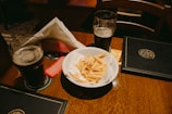 A wooden table features two glasses of dark beer, coaster mats beneath, and a white bowl filled with French fries. The table also holds a couple of menus and condiment packets beside the fries. There are napkins organized neatly in a holder.