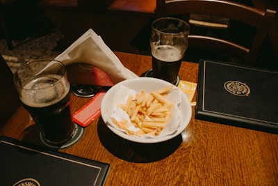 A wooden table features two glasses of dark beer, coaster mats beneath, and a white bowl filled with French fries. The table also holds a couple of menus and condiment packets beside the fries. There are napkins organized neatly in a holder.