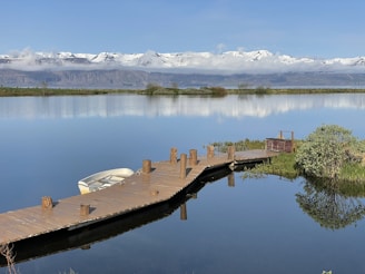 A wooden dock stretching into calm lake waters with a small rowboat tied up, framed by mountains.