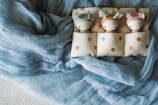 Three knit dolls with animal-like ears rest in a cozy fabric bed adorned with polka dots. The dolls are arranged side by side, each sporting a different pastel-colored outfit. Surrounding them is a soft, textured, blue fabric creating a serene and comforting atmosphere.