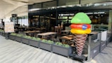An outdoor seating area of a restaurant named Nosolo Italia. There are several tables and chairs arranged neatly with plants as barriers. A large, colorful ice cream sculpture stands prominently near the restaurant entrance. The restaurant signage includes the words 'Pizzeria - Gelateria'. A menu board with images of food stands beside the seating area.