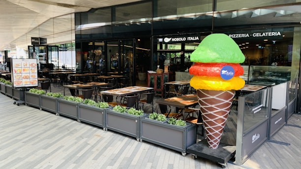An outdoor seating area of a restaurant named Nosolo Italia. There are several tables and chairs arranged neatly with plants as barriers. A large, colorful ice cream sculpture stands prominently near the restaurant entrance. The restaurant signage includes the words 'Pizzeria - Gelateria'. A menu board with images of food stands beside the seating area.