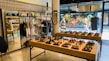 A stylish boutique featuring eyewear and clothing. Sunglasses are displayed on wooden tables, organized neatly in rows. Hanging racks in the background hold various garments, and shelves above display accessories. A mirror is placed on the left side, reflecting part of the store, and a person is browsing nearby. The storefront's large glass windows provide a view of people walking outside, with vibrant decorations in the window.