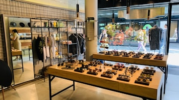 A stylish boutique featuring eyewear and clothing. Sunglasses are displayed on wooden tables, organized neatly in rows. Hanging racks in the background hold various garments, and shelves above display accessories. A mirror is placed on the left side, reflecting part of the store, and a person is browsing nearby. The storefront's large glass windows provide a view of people walking outside, with vibrant decorations in the window.