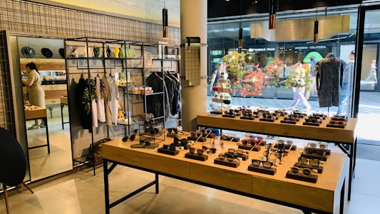 A stylish boutique featuring eyewear and clothing. Sunglasses are displayed on wooden tables, organized neatly in rows. Hanging racks in the background hold various garments, and shelves above display accessories. A mirror is placed on the left side, reflecting part of the store, and a person is browsing nearby. The storefront's large glass windows provide a view of people walking outside, with vibrant decorations in the window.