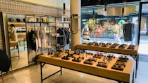 A stylish boutique featuring eyewear and clothing. Sunglasses are displayed on wooden tables, organized neatly in rows. Hanging racks in the background hold various garments, and shelves above display accessories. A mirror is placed on the left side, reflecting part of the store, and a person is browsing nearby. The storefront's large glass windows provide a view of people walking outside, with vibrant decorations in the window.