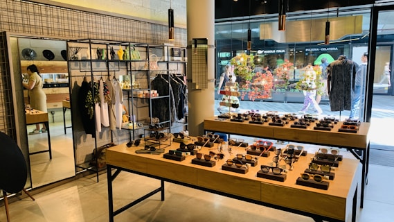 A stylish boutique featuring eyewear and clothing. Sunglasses are displayed on wooden tables, organized neatly in rows. Hanging racks in the background hold various garments, and shelves above display accessories. A mirror is placed on the left side, reflecting part of the store, and a person is browsing nearby. The storefront's large glass windows provide a view of people walking outside, with vibrant decorations in the window.