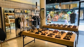 A stylish boutique featuring eyewear and clothing. Sunglasses are displayed on wooden tables, organized neatly in rows. Hanging racks in the background hold various garments, and shelves above display accessories. A mirror is placed on the left side, reflecting part of the store, and a person is browsing nearby. The storefront's large glass windows provide a view of people walking outside, with vibrant decorations in the window.