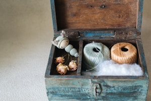 A vintage wooden box with sections containing dried roses, a small knitted doll, two spools of thread in mint green and beige, and a layer of fluffy cotton.