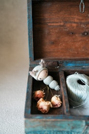 A wooden box with a rustic appearance contains a knitted toy resembling a small caterpillar, a spool of pale green thread, and three dried roses. The box features a weathered blue paint, adding a vintage charm to the scene.