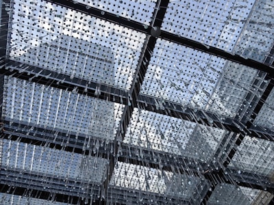 A modern architectural ceiling composed of a grid of glass squares, each adorned with numerous hanging glass droplets. The grid structure is supported by black metal frames, allowing natural light to penetrate through. The sky with scattered clouds is visible through the transparent ceiling.