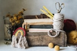 A rustic wooden box filled with various items such as books, a coiled rope, and a pair of scissors. There are dried yellow flowers in a jar on the left, balls of yarn, and a handmade rainbow decoration. A measuring tape is partially visible, wound up with one end sticking out.