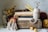 A rustic wooden box filled with various items such as books, a coiled rope, and a pair of scissors. There are dried yellow flowers in a jar on the left, balls of yarn, and a handmade rainbow decoration. A measuring tape is partially visible, wound up with one end sticking out.