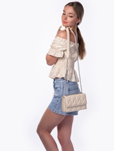 A woman with long hair styled in a ponytail is wearing an off-the-shoulder, smocked blouse with subtle floral patterns. She pairs it with a denim mini skirt and carries a quilted beige handbag over her shoulder. Her expression is serene as she looks down slightly with a soft smile.