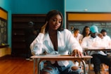 A young woman sitting at a school desk in a classroom, wearing a white blouse and ripped jeans, with a thoughtful expression. Several people are sitting at desks behind her, and there is a blackboard and bookshelves in the background.