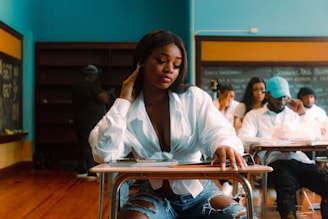 A young woman sitting at a school desk in a classroom, wearing a white blouse and ripped jeans, with a thoughtful expression. Several people are sitting at desks behind her, and there is a blackboard and bookshelves in the background.